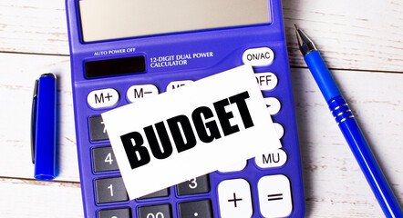 A large blue financial calculator, a blue pen and a blank card with the text BUDGET on a light...