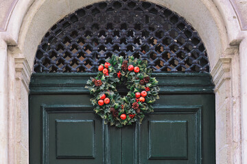 Traditional green and red Christmas wreath hanging on entrance door of house. Building facade decor during Christmas