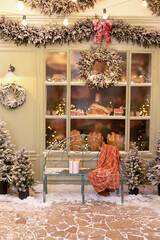 Xmas festive street with snow, bench and baking in showcase near a cafe bakery. Christmas decor: gift, toys, trees, garland fir branches, glowing light bulbs. Cozy outdoor cafe on Christmas eve.	