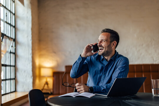 Male boss laughing while talking to someone over the phone, looking through the window, holding glasses.