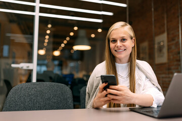 Portrait of a smiling girl, working online over the laptop while using a mobile phone.