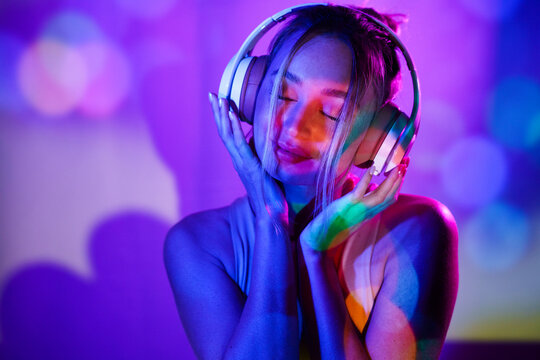 Smiling young woman with eyes closed listening to music through headphones in front of wall