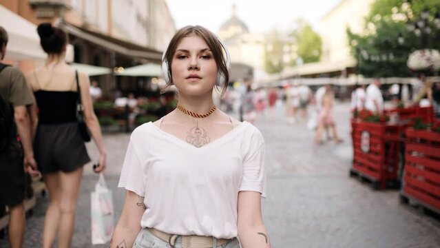 Outdoor summer lifestyle portrait of young pretty hipster woman in wireless headphones having fun, listening music and dancing on the street. Beautiful sexy woman with tattoo dancing on a street
