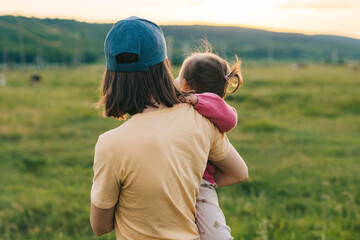 Back view of a mother wearing hat carrying the baby child in her arms, walking through the green field. Carefree childhood. People lifestyle. Summer vacation