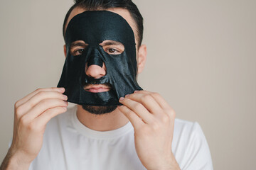 Portrait of a young man peeling off a black facial mask isolated over gray background. Health care. Beauty face. Beauty portrait. Beauty treatment. Aging care.