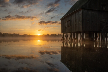 Fototapeta na wymiar Pfahlhaus am Teich bei Sonnenaufgang. Nebel über und Spiegelung im Wasser. Morgenstimmung