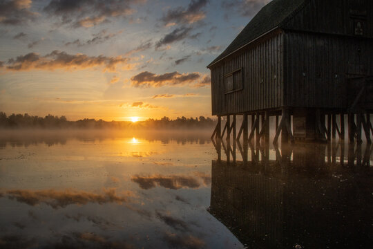 Pfahlhaus am Teich bei Sonnenaufgang. Nebel &uuml;ber und Spiegelung im Wasser. Morgenstimmung