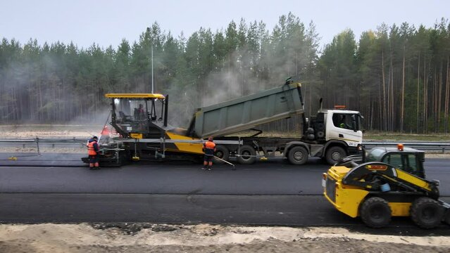 Road works. Laying new asphalt on an intercity highway. The truck pours the hot mix into the paver. Workers in overalls are watching the process. In the foreground is a small excavator. Roadbed repair