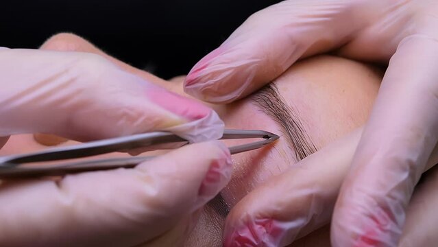 The master stretches the eyebrow with his fingers and removes unwanted hairs with tweezers. Cosmetic procedure permanent eyebrow makeup. Eyebrow tattooing.