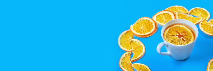 Circle of dried oranges with mug of tea on blue background. Banner.