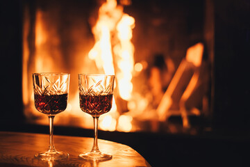 Two glasses of red wine on wooden table with cozy fireplace flame on the background.