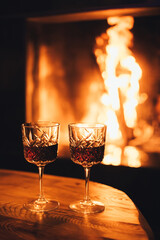Two glasses of red wine on wooden table with cozy fireplace flame on the background.