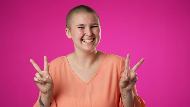 Portrait of happy gender fluid non binary young woman 20s smiling and giving peace or victory sign gesture with hands isolated on pink background.