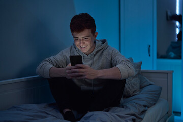Cheerful caucasian teenage boy using mobile phone while sitting at night in his room