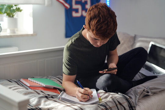 Caucasian teenager boy sitting on bed and studying using phone