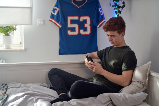 Caucasian teenage boy browsing phone while sitting on bed  in his bedroom