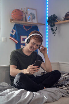 Caucasian teenage boy browsing phone with smile and wearing headphones while sitting on bed