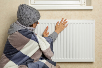 A frozen woman dressed in a blanket sits near a radiator in a cold room in winter.
