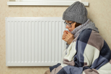 A woman in a cold room, wrapped in a blanket, warms herself near the heating radiator.