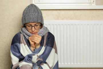 A frozen woman dressed in a blanket sits near a radiator in a cold room in winter.