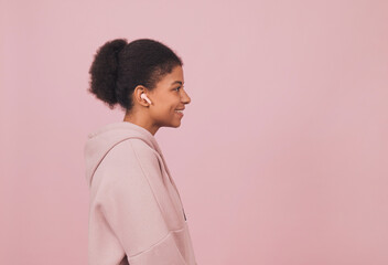 Obraz na płótnie Canvas Happy young black woman in wireless headphones. Side-profile portrait of african american girl in pink hoodie listening music against pink backdrop