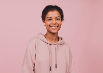 Happy smiling black girl is listening music. Young pretty woman in hoodie with wireless headphones against pink backdrop. Close up portrait in minimal style