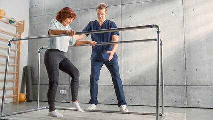 Fototapeta premium Physical Therapy Center: Portrait of Strong Senior Woman with Leg Injury Successfully Walks Holding Parallel Bars. Physiotherapist, Rehabilitation Doctor, Trauma Prevention Therapy Hospital.