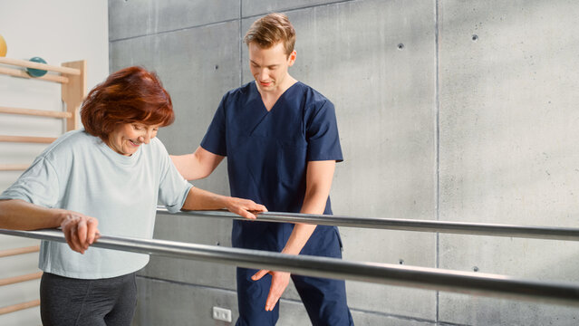 Portrait of Strong Senior Woman with Leg Injury Successfully Walks Holding Parallel Bars. Professional Physiotherapist Assists, Helps, Trains Disabled Person Do Rehabilitative Physiotherapy.