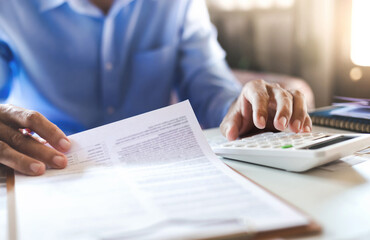 Asian businessman on desk in office using calculator to calculate life insurance benefits or expenses from statement and financial report,  Accountant working in the office, close-up view