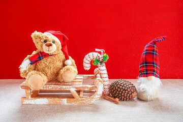 Holzschlitten mit Teddybär, Wichtel, Zuckerstange und Tannenzapfen vor einem roten Hintergrund