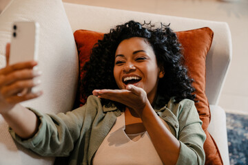 Young african woman blowing air kiss while using mobile phone in cozy apartment