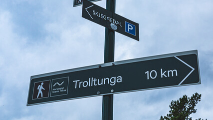 A dark green touristic directional sign on a hiking trail in Norway against the sky - Trolltunga 10 kilometers. The beginning of the pathway to Troll's Tongue. 