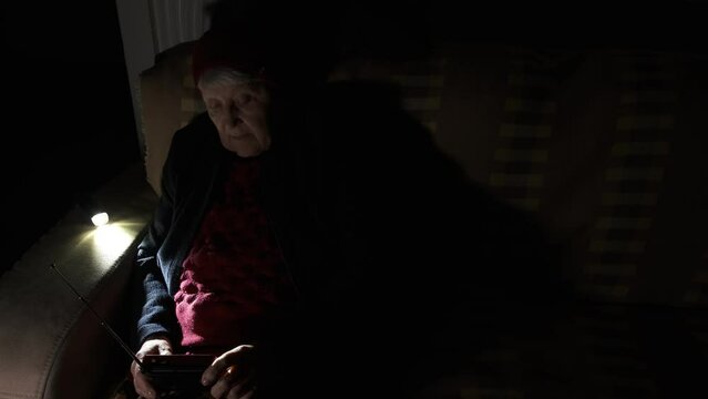 Ukraine. Grandmother holds radio in hands sitting in darkness blackout during russian terroristic attacks. Senior woman in winter clothes in darkness on sofa with hand radio receiver listening news.