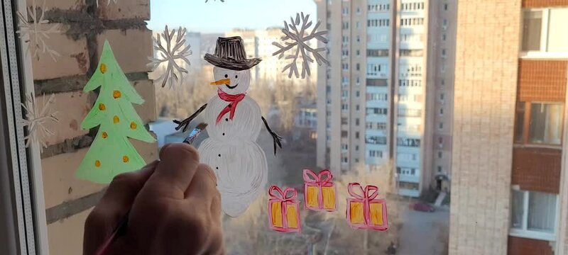 Person painting snowman, christmas tree and snowflakes on the window glass. Winter picture on the window.