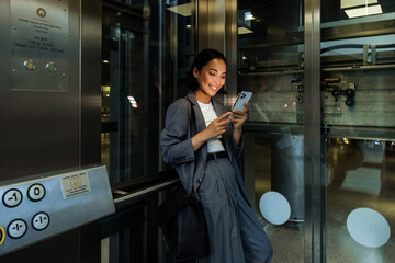 Young asian woman using smart phone while standing in elevator - obrazy, fototapety, plakaty