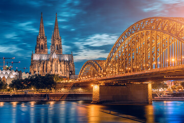 Scenic view of illuminated bridge over Rhine river whith trains and tourists passing by and the Cologne Cathedral in the evening at the blue hour.