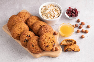 On a gray table are oatmeal cookies, honey, hazelnuts, oatmeal and cranberries in a plate.