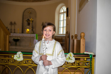  a handsome smiling boy in white clothes with a church candle stands in a Catholic church after...