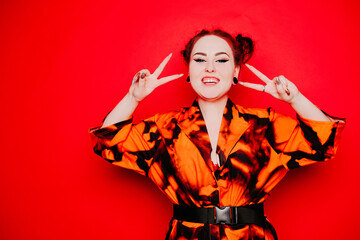 Beautiful girl on a red background. Red hair and tunnels in the ears, red dress and lips. Anime poses. The girl with eyeliner and two buns on her head grimaces, smiles, has fun. Peace gesture.