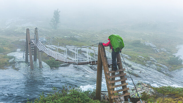 A woman hiker with a large green backpack crosses a stormy fast-moving mountain river in Norway on a suspension wooden bridge in rain and fog. Dangerous extreme river crossing. Climbing steep steps.