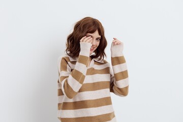 a sweet, pleasant woman with a short haircut stands on a light background in a stylish beige, striped sweater, and raises her hands to her face. Horizontal studio photography with empty space