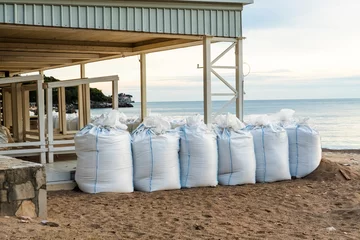 Foto op Canvas There are many large white sandbags on the beach for flood protection. Sandbags to protect buildings and coastal structures to protect against tsunamis and floods. water protection structures. © Григорий Галасюк