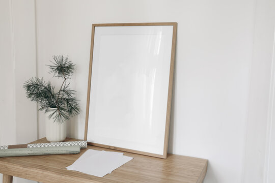 Christmas interior still life. Blank vertical wooden picture frame mockup on wooden table, desk. Pine tree branches in vase. Empty paper sheets, gift wrapping paper on desk. White wall background.