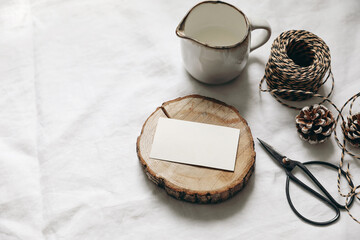 Christmas table setting. Winter breakfast composition. Blank business card mockup on wooden board. Milk pitcher, rope, pine cones and vintage black scissors on white linen. Seelective focus, top view.