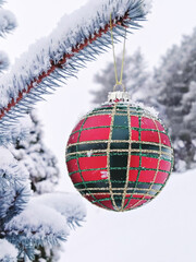 Red and green checkered big Christmas ball hanging on snow-covered spruce tree branch outside