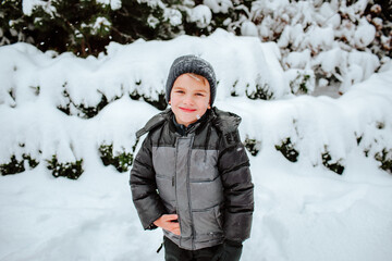 Portrait of cute young boy in grey winter jacket and hat against snowed winter garden background.