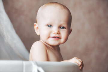 Head of little kid looking at camera from his infant cot. Happy hairless adorable baby with beauty blue eyes interested in white round crib. Cheerful child playing in sunny bedroom. Copy text space - obrazy, fototapety, plakaty