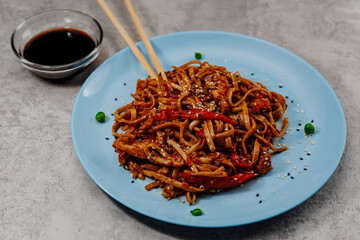 Spicy udon with chicken on a blue plate and soy sauce in a glass bowl on a gray background.