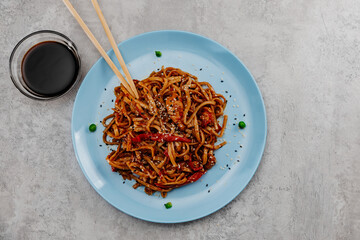 Spicy udon with chicken on a blue plate and soy sauce in a glass bowl on a gray background
