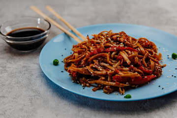 Spicy udon with chicken on a blue plate and soy sauce in a glass bowl on a gray background.